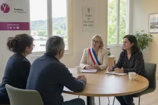 Three people in a sunlit municipal office engaged in calm discussion around a table, with a mediator and an official guiding the conversation.