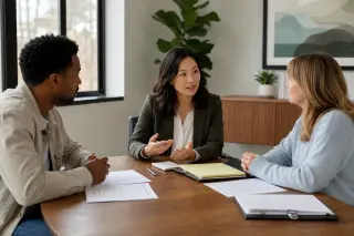 Three people, including a mediator, have a calm discussion around a table in a sunlit, modern meeting room, with documents and notes in front of them.